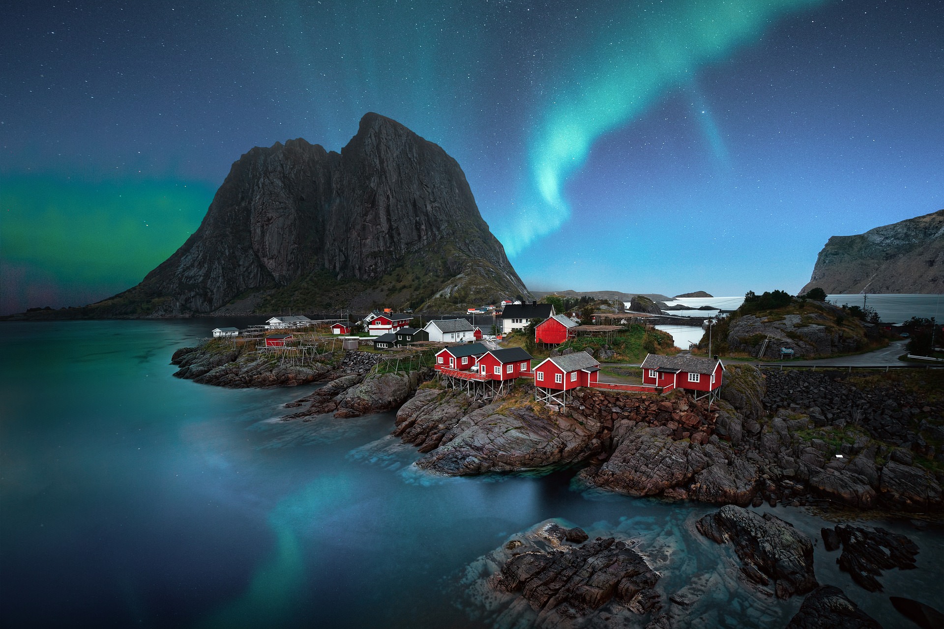 Vivid turquoise and green aurora swirling above traditional red rorbuer cabins in Hamnøy, Lofoten Islands