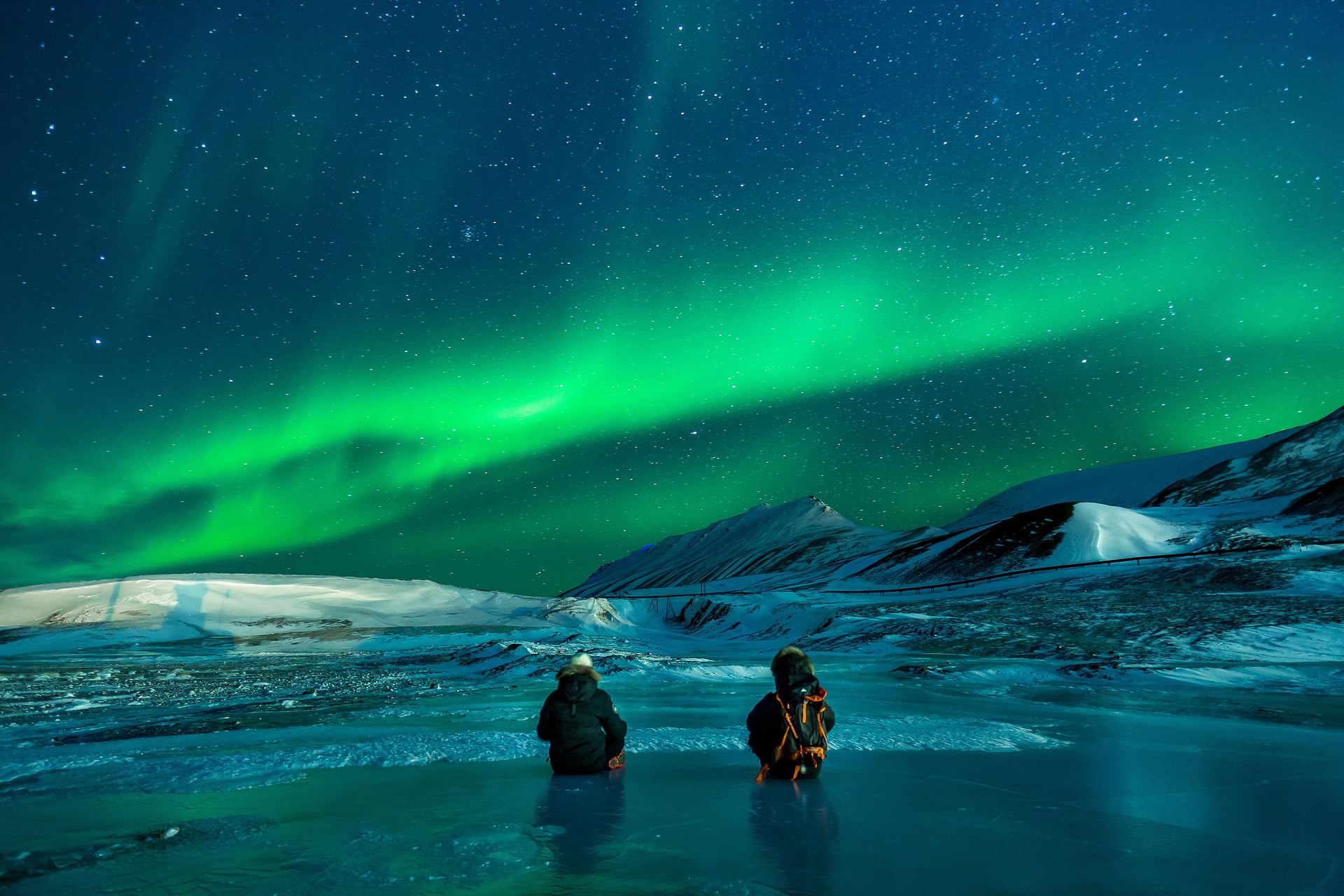 Couple sitting on frozen sea ice watching powerful green Northern Lights explosion in northern Norway