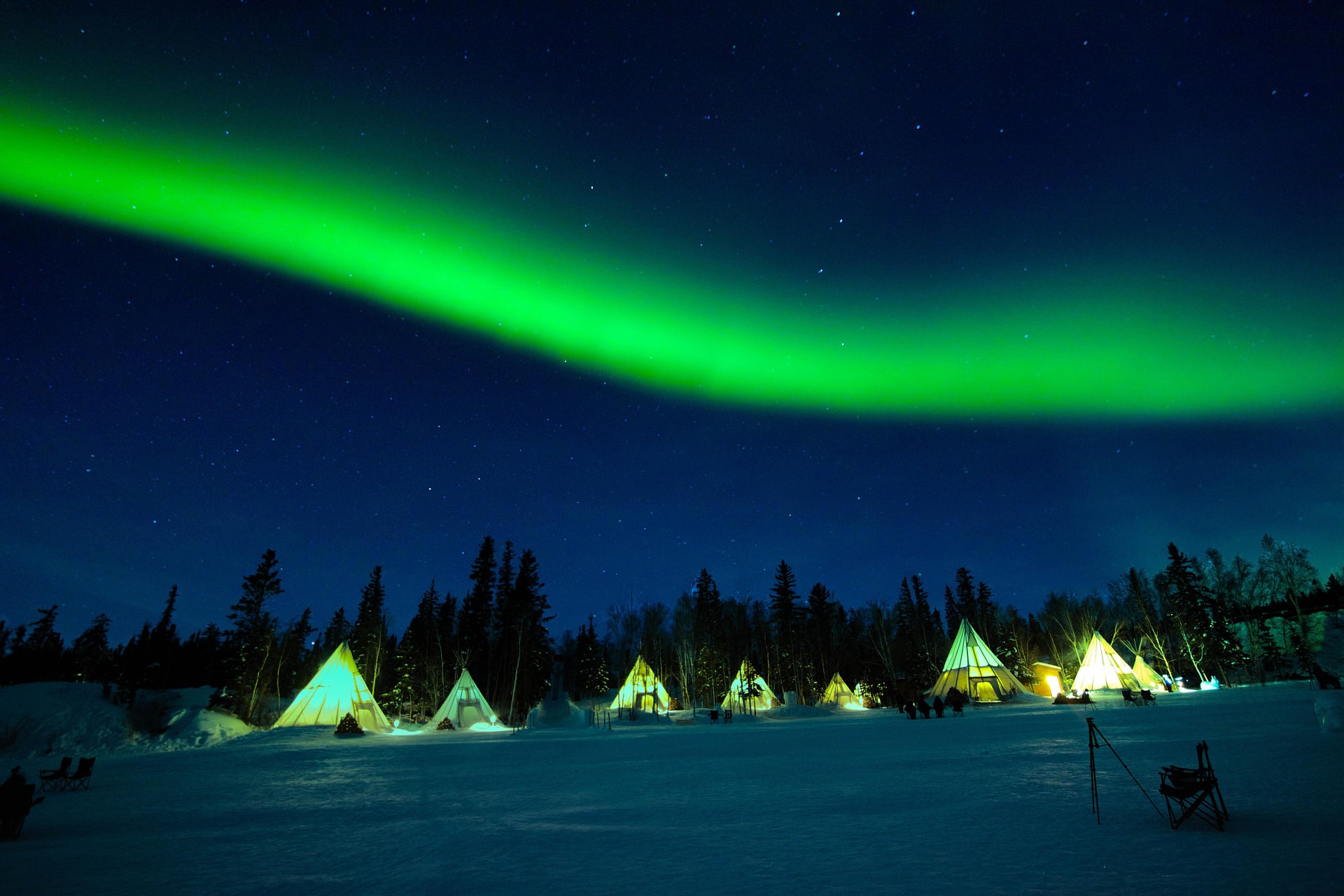 Bright green Northern Lights arching over illuminated Sami lavvu tents on a frozen lake in Lapland